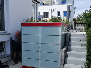 . The image depicts a modern, sleek outdoor storage unit placed next to a house. It features a minimalist design with frosted glass panels and a red accent at the top. There are stairs on the right side, and a person is partially visible on the left. The environment is clean and well-maintained, suggesting a residential area.