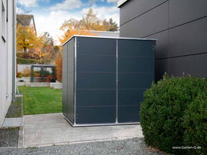 . The image shows a modern, sleek outdoor storage shed placed on a paved area next to a building. The shed has a rectangular shape with a dark finish and metallic edges. To the right, there's a bush, and behind the shed is a lawn leading to a landscaped area with trees in the background. The image also contains the URL "www.Garten-Q.de" at the bottom right corner.
