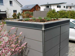 . Ein modernes Gartenhaus mit bepflanztem Dach, daneben blühende Pflanzen und Häuser im Hintergrund.