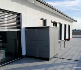 . The image shows a modern outdoor space with a flat-roofed white building. There is a large glass door with closed blinds on the side, and a sleek, gray storage cabinet placed against the wall. The area is paved with tiles, and there is a table and some potted plants on the patio. The setting appears to be on a rooftop or balcony, offering a clear sky view in the background.
