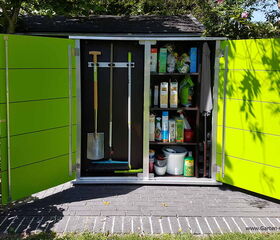 . The image shows an outdoor storage cabinet with open doors, revealing various gardening tools and supplies. On the left side, there are gardening tools like a shovel, spade, and broom hanging on hooks. The right side has shelves filled with various containers, bottles, and boxes probably containing fertilizers, plant food, or other garden-related products. The cabinet is bright green and situated on a patio with greenery around it.