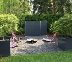 . The image shows an outdoor area with a setting that includes the following elements:

1. Two modern lounge chairs with colorful patterns.
2. A metal fire pit at the center of the chairs.
3. Two large square planters with small trees.
4. A gravel surface covering the area.
5. A dark-colored structure, possibly a shed or storage unit, in the background.
6. Lush greenery and trees surrounding the area, providing a natural backdrop.
7. A cat standing near one of the lounge chairs.

This arrangement appears to be a well-designed outdoor relaxation or lounge space.