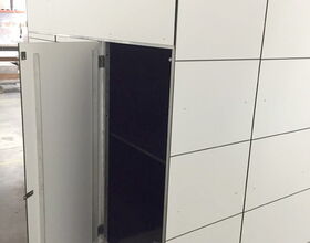 . The image shows a white storage unit or cabinet with a grid pattern on its exterior, and one of its doors is open. The interior appears dark. The unit is located in what seems to be an industrial or warehouse setting.