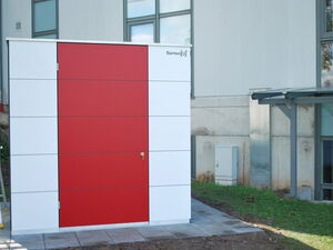 . The image shows a large, rectangular storage unit with a red front panel and white side panels. It's placed outside a building. The unit has a door with a lock, and there's some paving and grass around it. The design looks modern and minimalistic, and you can see part of the building, including windows and a small roof, in the background.