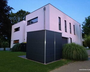 . The image shows a modern, minimalist two-story house with a flat roof and several narrow windows. In front of the house, there's a tall, dark-colored structure, possibly serving as a storage unit. The property is surrounded by grass and some bushes. Trees are visible in the background, and the sky is clear.