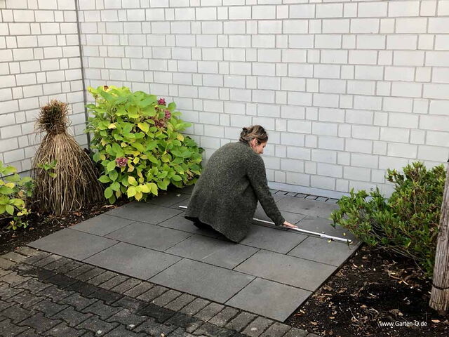 Eine Frau misst auf einer Terrasse mit Fliesen, umgeben von Pflanzen, an einer weißen Ziegelwand.