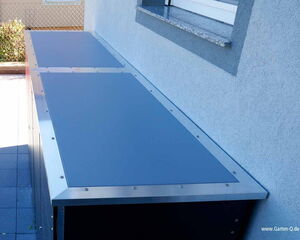 The image shows a large, rectangular outdoor storage cabinet with a flat, metal top. It is mounted against a wall. The storage unit appears to be robust and weather-resistant, possibly used for storing garden tools or equipment. There are screws or rivets around the edges of the top panel.