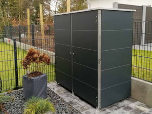 . The image shows a neat outdoor area featuring a modern, dark-colored storage unit. It is placed on a paved surface surrounded by a landscaped garden. There's a planter with dried plants and another with green grass, set on a bed of gravel. The area is enclosed by a fence and nearby is a house with visible roofing and walls.