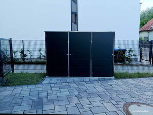 . The image shows a modern outdoor storage shed, likely made of metal or composite materials. It is situated on a paved area with a fence and some greenery in the background. The sleek design suggests it is used for storing garden tools or equipment.