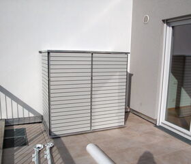 This image shows a patio or balcony area with a horizontal-slatted storage unit against a wall. There are some railing bars casting shadows on the ground, a grate-like fixture embedded in the floor, and part of a sliding glass door is visible to the right. Some long, white objects, possibly pipes or rods, are on the floor in the foreground.