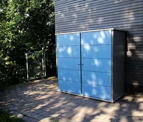 . Ein blauer Gartenschrank steht auf einer gepflasterten Fläche neben einem Gebäude, umgeben von Bäumen.