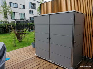 . Ein grauer Gartenschrank auf einer Terrasse, daneben ein Garten mit Trampolin und mehrstöckigem Gebäude im Hintergrund.
