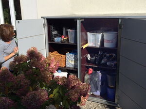 . Ein offener Schrank im Garten mit verschiedenen Haushaltsgegenständen. Eine Person schaut hinein.