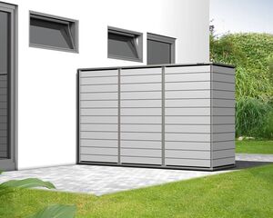 The image shows a modern outdoor storage unit or shed placed next to a building. It is designed with horizontal panels and is positioned on a paved area. The wall of the building has two small windows, and there is greenery, including grass and plants, in the surrounding area.