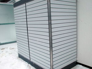 . The image shows a large, outdoor cabinet or storage unit placed against a building wall. The cabinet has a sleek design with horizontal slats. It's surrounded by snow, indicating a cold or winter setting. The area appears to be an exterior space, possibly part of a residence or commercial building.