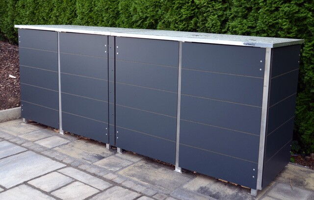 . The image shows a sleek, modern outdoor storage unit with three compartments, likely made of metal, situated on a tiled patio. It has a flat top and is positioned against a green hedge.