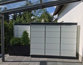 . The image depicts a modern outdoor setting with several features:

1. A gray tiled patio or terrace area.
2. Two large, light gray cabinets or storage units.
3. A planter with green bushes to the left.
4. A glass or transparent roof or awning structure above.
5. Trees and greenery in the background alongside a white building wall.