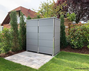 The image shows a modern outdoor storage shed with a sleek, gray design. It is situated on a tiled patio area in a garden, surrounded by trees and shrubs. There is a house in the background with a red-tiled roof and a satellite dish.