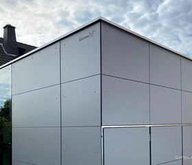 Modernes Gartenhaus mit grauen Paneelen, befindet sich neben einem Gebäude mit Schornstein, Himmel im Hintergrund.