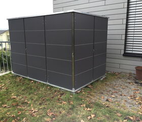. The image shows a metal storage shed placed on a patch of grass next to a building. There are fallen leaves scattered on the grass, and a brown empty pot is visible near the building. The shed has smooth, dark-colored panels and is located near a metal railing.