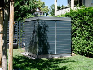 . The image shows a modern, rectangular outdoor shed with horizontal slats and a sleek design. It's situated on a grassy area, surrounded by trees and shrubs. The shed appears to be made of metal or a similar material and is likely used for storage in a garden or backyard setting.