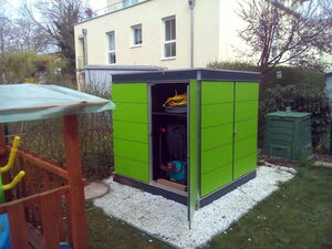 . The image shows a small, green garden shed with an open door, revealing some equipment inside. The shed is placed on a bed of white gravel. In the background, there is a building with windows, some trees, and a compost bin. To the left, part of a wooden structure with a transparent roof is visible. The scene appears to be set in a backyard or garden.