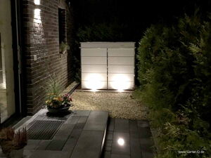 . Ein beleuchteter Außenbereich mit Pflanzgefäß und weißem Schrank neben einer Ziegelmauer und Hecke.
