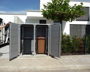 . The image shows a storage unit or shed with open doors, housing two waste bins—one black and one brown. This setup is located outside, near a building, with some greenery and a tree in the background.