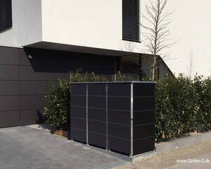Here's a breakdown of the image features in an array format:

1. **Building**: Modern structure with a minimalist design.
2. **Wall Cladding**: Dark panels on part of the exterior.
3. **Window**: Rectangular, with a louvered design.
4. **Bushes**: Green plants along the side of the building.
5. **Tree**: Leafless, possibly in a dormant season.
6. **Storage Unit**: Black, rectangular, metallic structure.
7. **Pavement**: Grey, stone-like tiles.
8. **Website Watermark**: URL text "www.Garten-Q.de" at the bottom right.