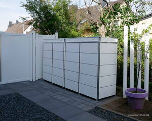 The image shows a series of modern outdoor storage units arranged neatly against a white fence. The units have a sleek design with horizontal lines. There is a purple pot with a plant next to the storage units. The setting is outdoors, and there's a paved area with tiles and gravel. Trees and a building are visible in the background.