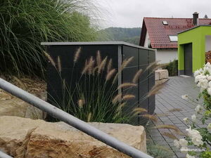 . The image shows a modern outdoor storage unit with a sleek design. It's situated in a garden setting with ornamental grasses and white flowers around it. In the background, there are buildings featuring red-tiled roofs. The area has a paved pathway leading to the structures.