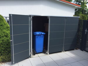 . The image shows an outdoor storage enclosure with one door open, revealing a blue wheeled bin inside. The enclosure is situated on a tiled patio area, with a hedge nearby. The structure is made of dark-colored panels and has an open design on one side.