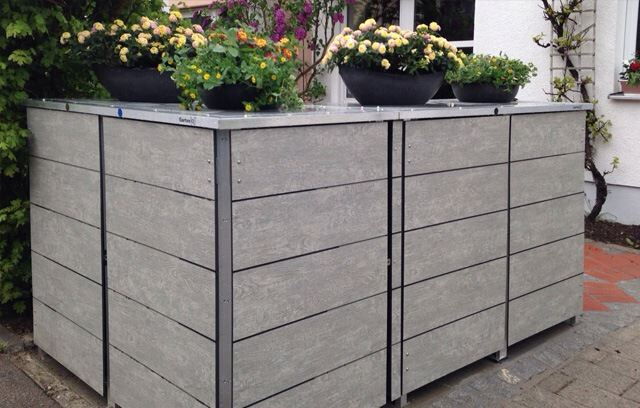 . The image shows a large outdoor storage unit with a wooden or composite finish, likely used for trash bins or recycling containers. On top of the unit, there are planters with blooming flowers, adding a decorative touch to the otherwise utilitarian structure. The setting is possibly the side or front of a house, with some greenery and brick paving in view.