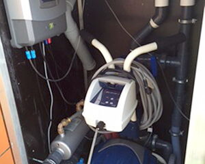 The image shows a pool cleaning device setup, featuring equipment such as a robotic pool cleaner with some attached hoses and technological components. It is housed in a compartment, with control units and piping visible. The setup appears organized for maintaining pool cleanliness.