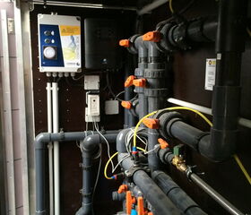 The image shows an arrangement of plumbing and electrical components. There are several pipes, valves, and connectors organized in a network, likely for controlling water flow. The valves have orange handles. There is also a control box on the left side with buttons and indicators, connected to the system, possibly for managing the operation of the system. Yellow wires and tubing are visible alongside the plumbing components. This setup is typical in mechanical or utility rooms for managing fluid systems.