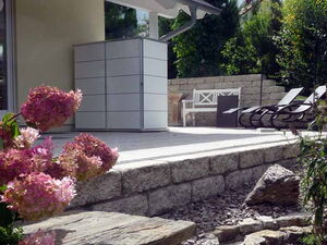 . The image depicts an outdoor patio area. Key elements in the scene include:

- A tiled house roof and part of the house wall.
- Outdoor seating with lounge chairs positioned on a wooden or composite deck.
- Stone steps and a stone retaining wall.
- Pink flowering plants and green bushes.
- Open skies with some clouds in the background.

It appears to be a well-maintained, landscaped patio or garden area.