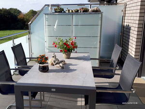 . Ein Balkon mit Tisch, vier Stühlen, Pflanzen und Dekoration vor einer Sichtschutzwand aus Glas.