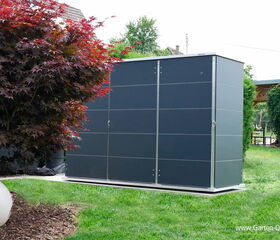 . The image shows a modern garden storage unit with a sleek, dark exterior. It's situated in a well-maintained garden with a lawn and some ornamental plants, including a tree with reddish leaves on the left. There is also a small white spherical object on the ground near the tree.