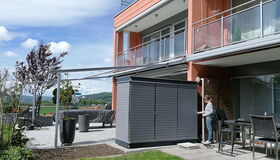 . Moderne Terrasse mit Pflanzen, Möbeln und Schuppen vor einem zweistöckigen Gebäude; eine Person steht daneben.