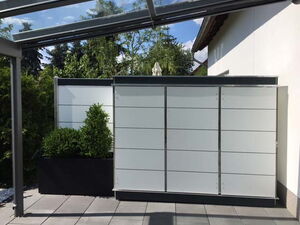 . Terrasse mit Sichtschutzwänden aus Glas und Pflanzenkübel, überdacht mit Glasdach, umgeben von Bäumen.
