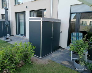 . Garten, Terrasse mit Schrank, Liegestuhl, Sonnenschirm und Pflanzen vor einem modernen Gebäude.