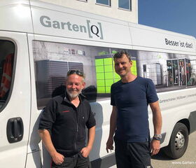 . Zwei Männer vor einem Lieferwagen von Garten[Q] mit Werbung für Gartenschränke, Müllboxen und Unterstände.