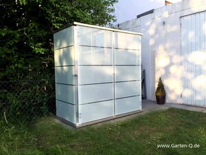. The image shows a modern, rectangular outdoor storage shed located in a garden area. It appears to be made of metal or a similar material, with horizontal lines and a sleek design. The shed is placed near a fence, surrounded by greenery, and there's a concrete pathway beside it. A potted plant is visible near a wall to the right.