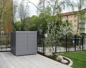 . Ein graues Gartenhäuschen steht auf einem gepflasterten Hof, mit Bäumen und Wohngebäuden im Hintergrund.