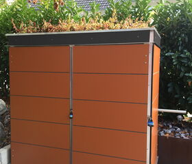 The image depicts a storage shed with a green roof. The shed has orange paneling, a metal frame, and is secured with locks. There is vegetation growing on the roof, and a leafy hedge or bushes are in the background. A house with a tiled roof is also visible behind the shed.