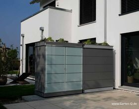 The image depicts a modern garden storage unit or shed situated outside a house. It is sleek, with a combination of frosted glass and metal panels. The unit is placed on a patio area next to a contemporary building. There are plants visible on top of the unit and surrounding the area.