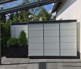 . The image shows a modern patio area with a sleek design. There is a planter with greenery on the left, and a large privacy screen made of frosted or opaque panels. The patio has tiled flooring, and there's a partial view of a structure with a glass roof on the left side. The area is adjacent to a house wall.