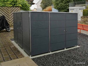 . The image shows a large, outdoor storage unit or shed, likely made of metal or a similar material. It consists of multiple compartments with doors that have handles. The structure is placed on a gravel surface, surrounded by some paving stones. There’s a motorcycle covered nearby and a red bench in the background. The area is surrounded by greenery and some other structures are visible in the distance.