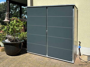 . Ein graues Gartenschrank mit Topfpflanze daneben, steht auf einer Terrasse neben einer Hauswand.
