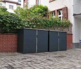 Zwei graue Mülltonnenboxen stehen nebeneinander auf einem gepflasterten Hof nahe einer roten Backsteinmauer.